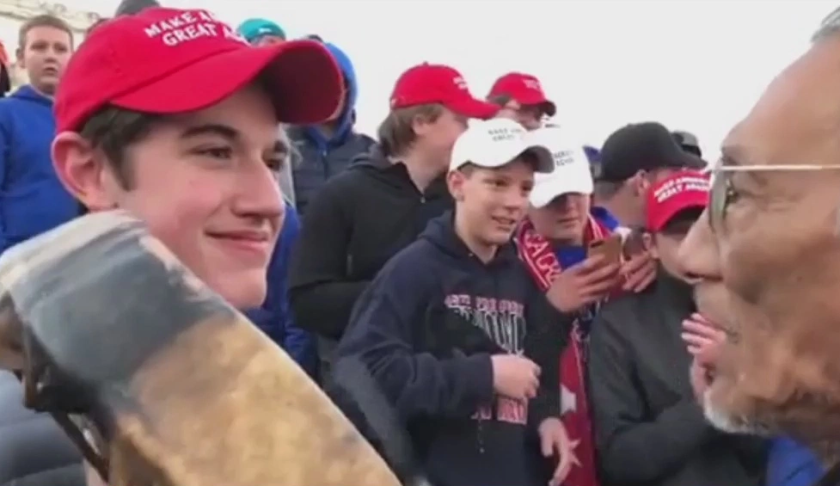 A teenager wearing a "Make America Great Again" hat stands in front of an elderly Native American singing and playing a drum in Washington. The Roman Catholic Diocese of Covington in Kentucky is looking into this and other videos that show youths, possibly from the diocese's all-male Covington Catholic High School, mocking Native Americans at a rally in Washington. Photo via CBS News.