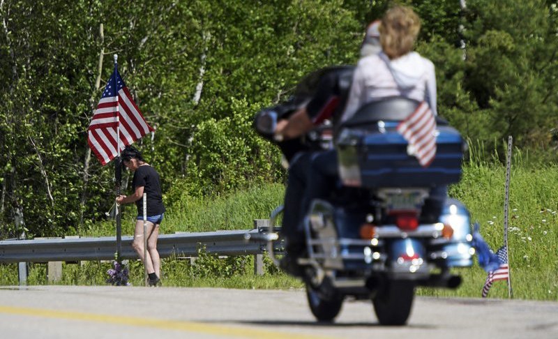 A motorcycle passes as a woman leaves flowers at the scene of a fatal accident on Route 2 in Randolph, N.H., Saturday, June 22, 2019. Investigators pleaded Saturday for members of the public to come forward with information that could help them determine why a pickup truck hauling a trailer collided with a group of motorcycles on a rural highway. (Paul Hayes/Caledonian-Record via AP)